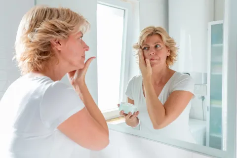 Skincare en la menopausia: cómo tener una piel radiante en esta etapa de la vida Una mujer mirándose al espejo mientras se aplica una crema