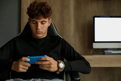 Ludopatía en adolescentes: testimonios en primera persona Adolescente con celular en la mano y computadora de fondo.