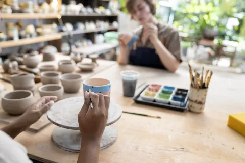 El boom de las experiencias: 5 planes creativos para hacer con amigas Manos de mujer decorando una pieza de cerámica y una mujer en segundo plano pintando una cerámica.