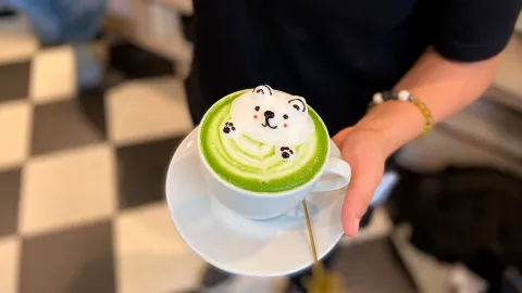 Salir a merendar con los chicos en vacaciones: 5 cafeterías con juegos que les van a encantar Plano medio de una persona sosteniendo un plato con una taza de café. En ella se encuentra un café de color verde con el relieve de espuma con forma de oso tipo kawai