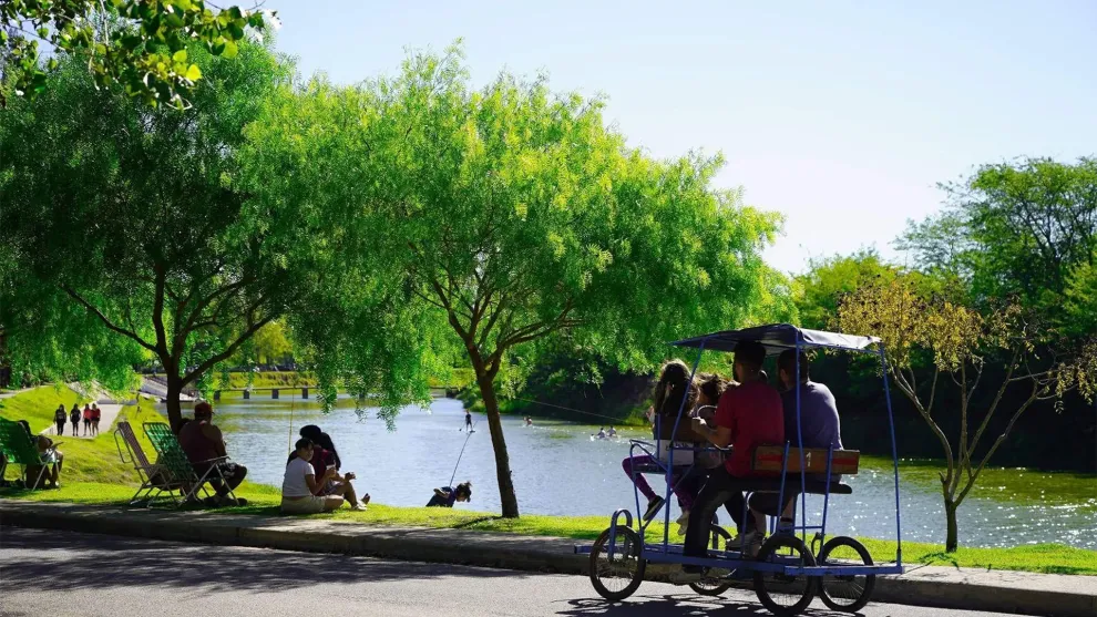 Termas de General Belgrano, un lugar donde se ve un espejo de agua y un espacio verde con gente descansando. En primer plano, un carrito de paseo con cuatro personas pedaleando.